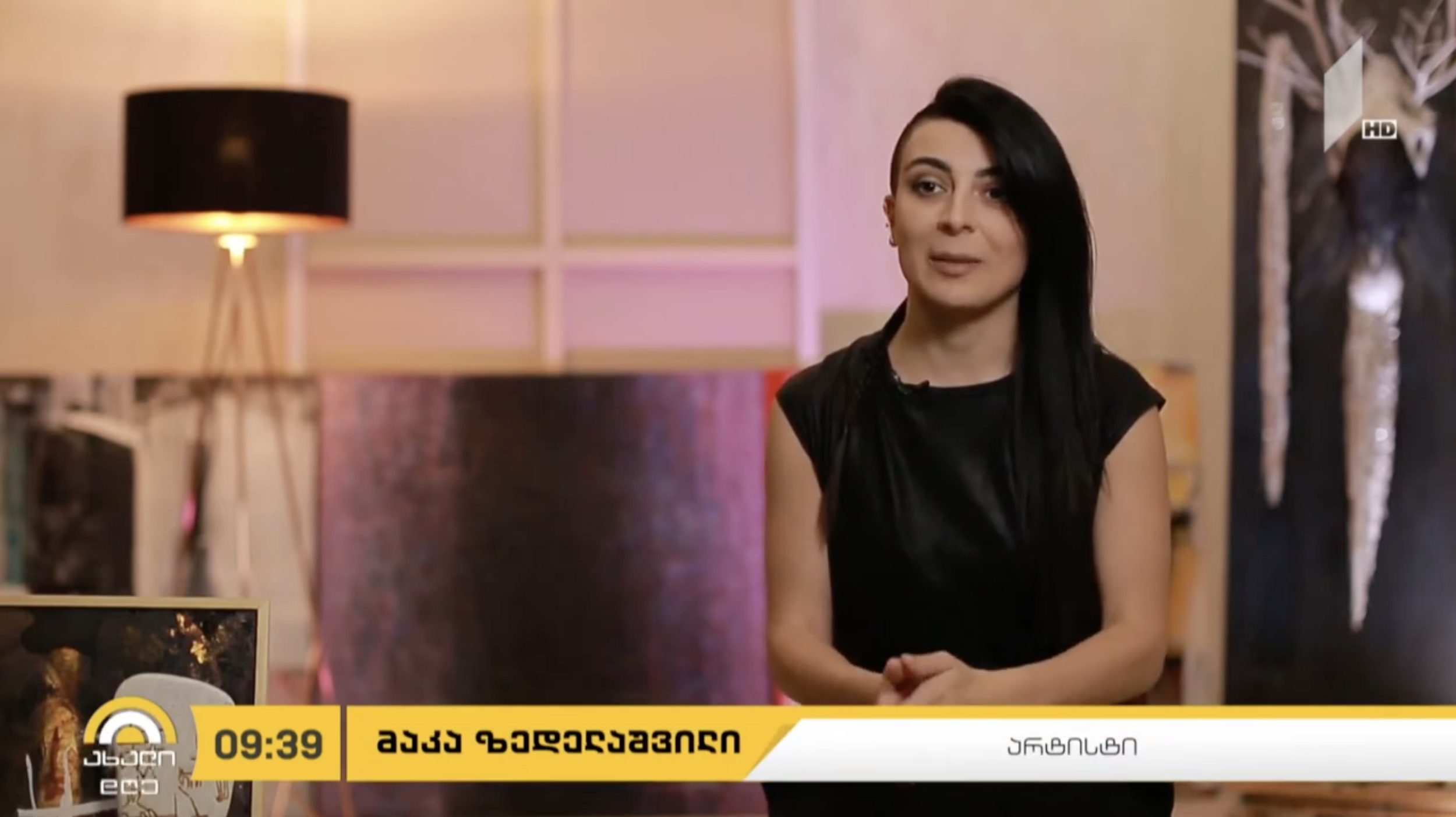 A woman with black hair styled in an asymmetrical undercut, wearing a black sleeveless top, standing indoors in front of a decorative background with a lamp and artwork. A yellow news banner with Georgian text is at the bottom of the image.