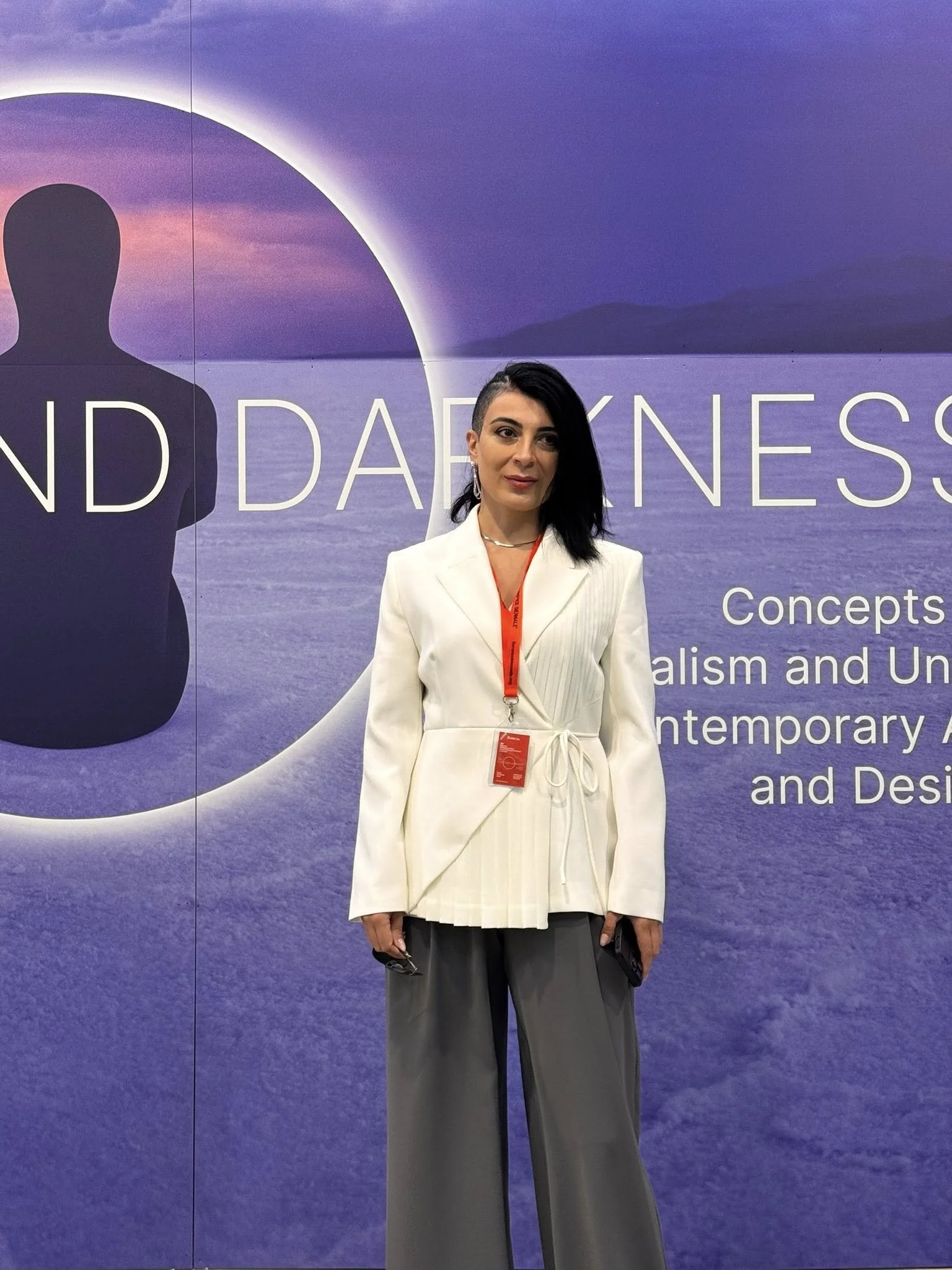 A woman with dark hair, one side shaved, wearing a cream-colored blazer and gray pants, standing in front of a large backdrop with ocean and sky imagery, and partial text about art and contemporary design.