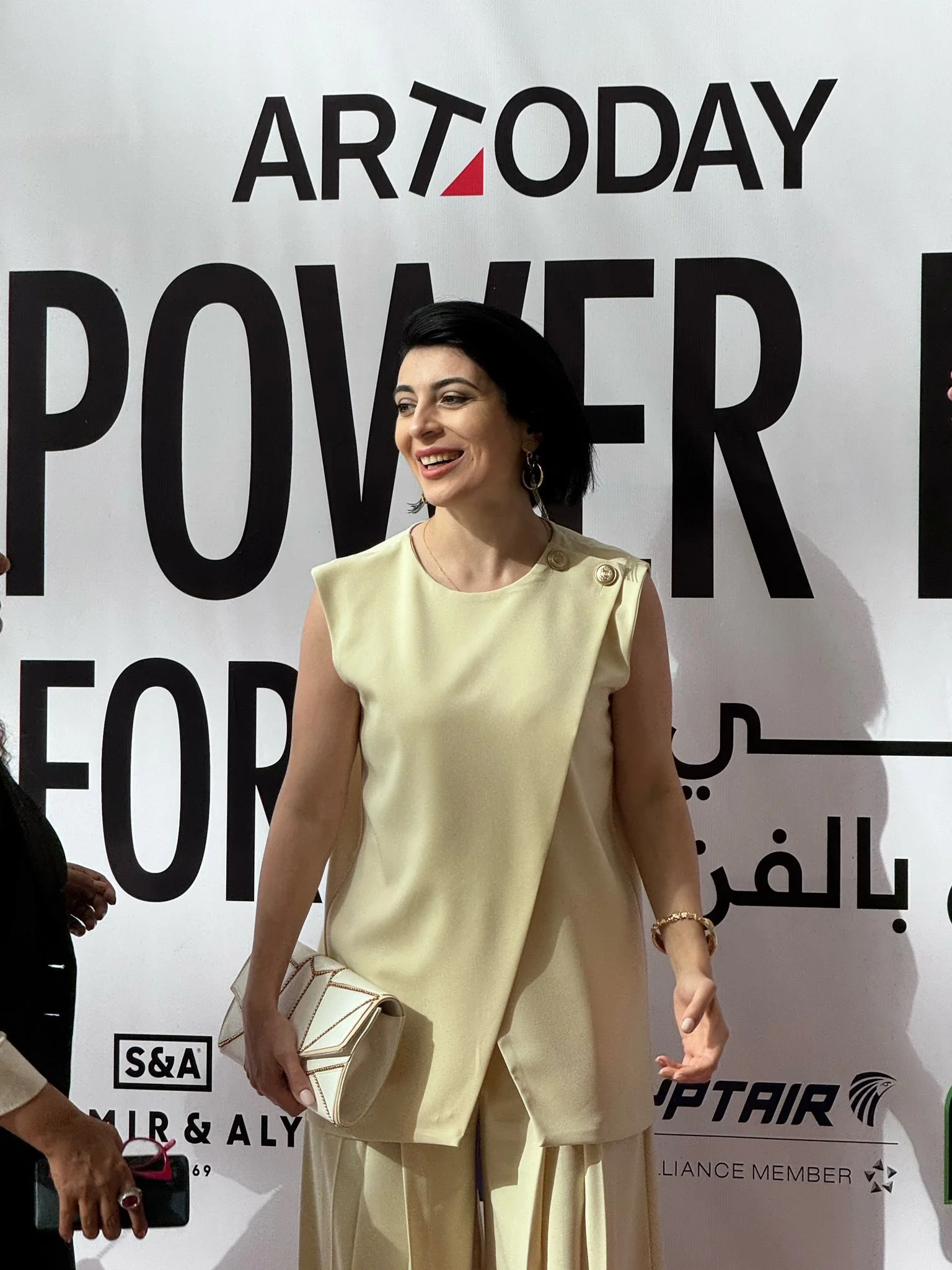 Woman wearing a cream-colored outfit holding a matching handbag, standing in front of a white background with large black text and logos.