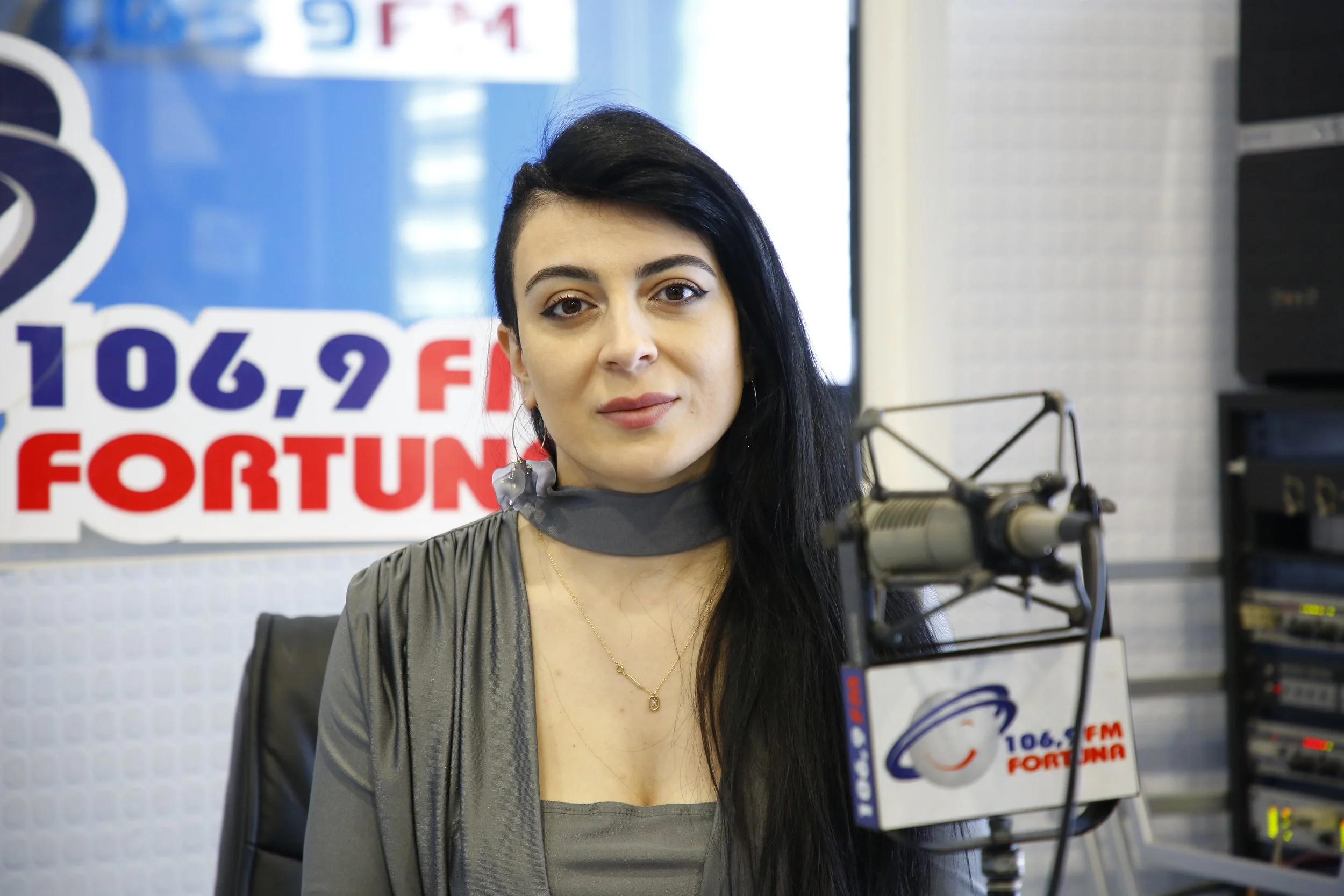 A woman with black hair and a side shave haircut sitting in a radio studio with a microphone. There is a FORTUNA radio station sign behind her. The studio has a panel with various audio equipment.