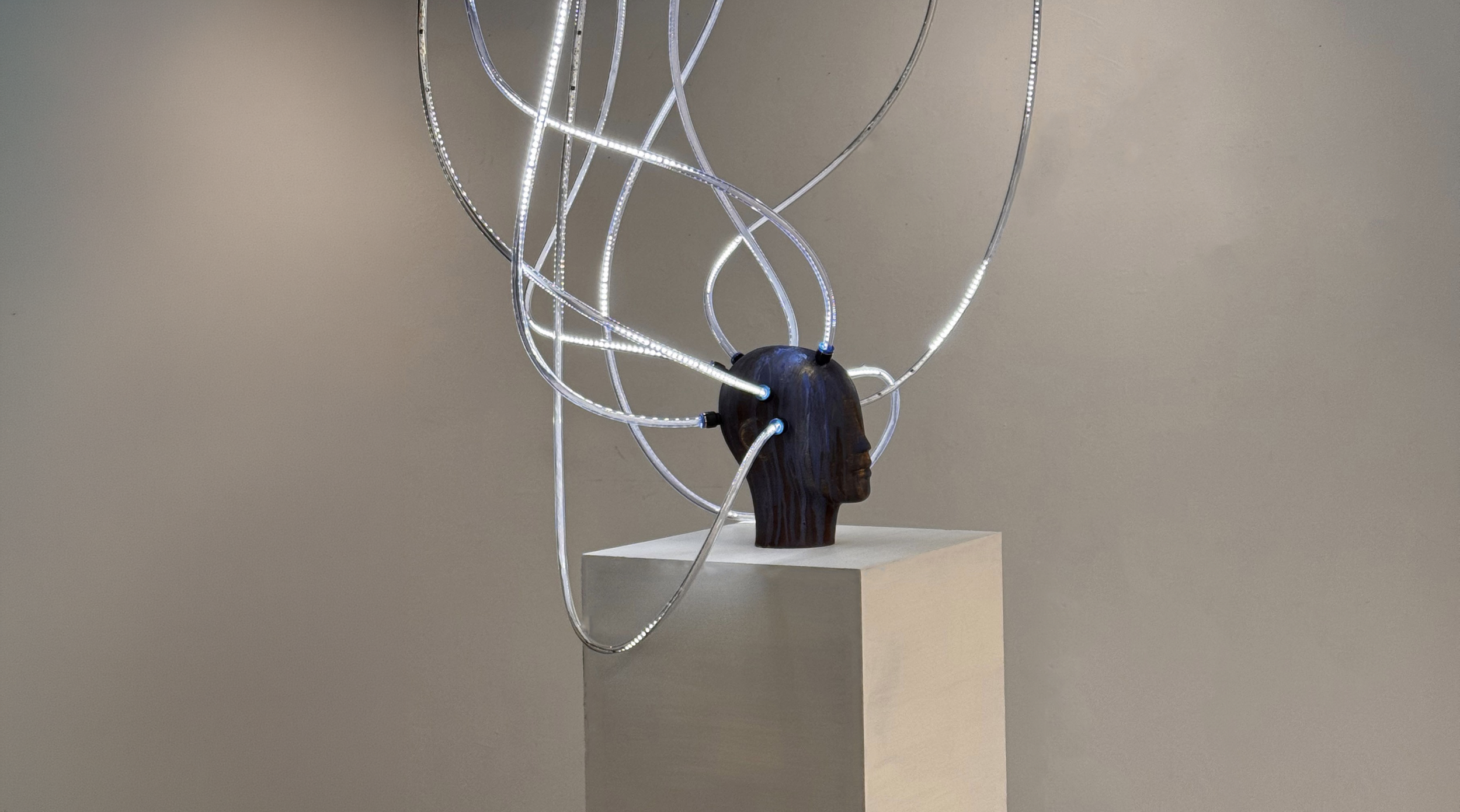 Art installation featuring a human head sculpture with illuminated loops emerging from it, displayed on a pedestal against a plain wall.