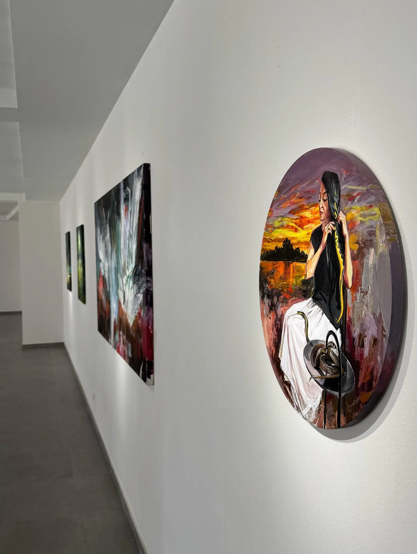Art gallery with multiple paintings, including a circular portrait of a woman with long black hair sitting outdoors at sunset.