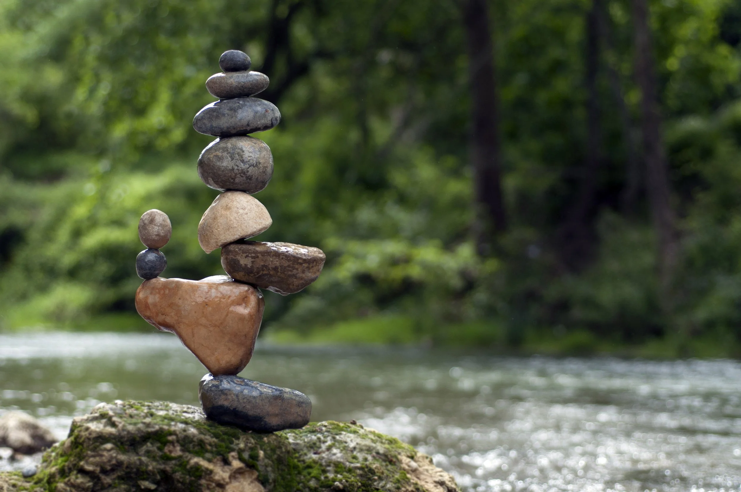 Stacked river rocks on mossy rock beside flowing river with green trees in background.