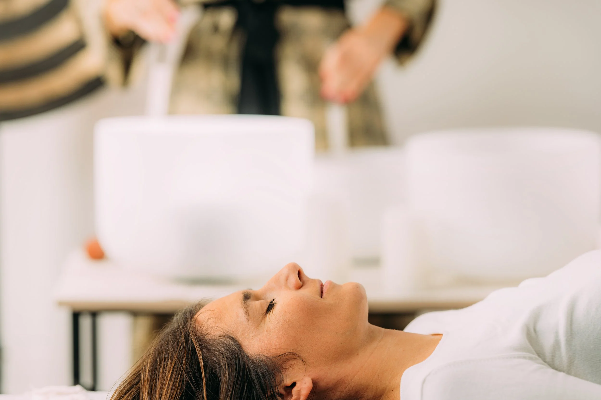 A woman lying down in a relaxed state with her eyes closed, receiving a sound healing session with crystal singing bowls.