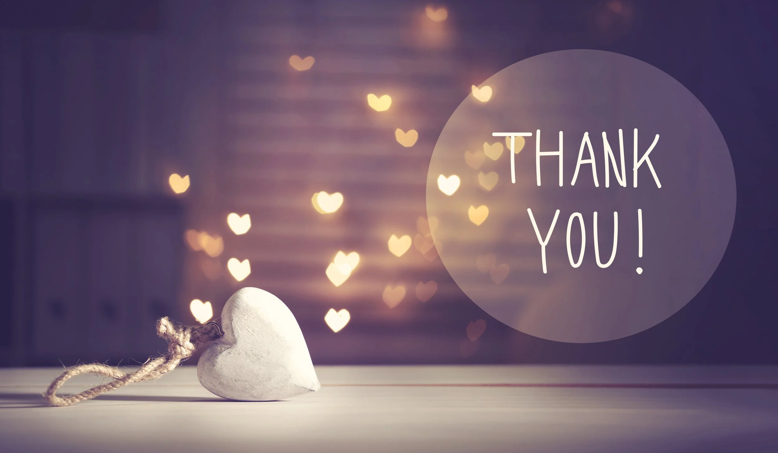A white heart-shaped ornament with a twine bow on a light surface, with blurry heart-shaped lights in the background and a semi-transparent circle containing the words 'Thank You!'