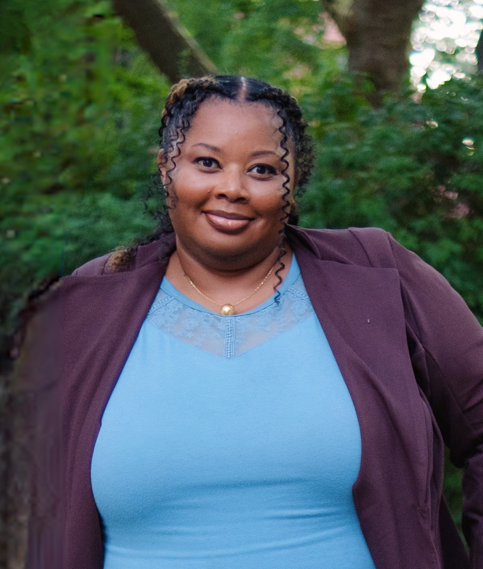 A woman smiling outdoors wearing a blue top, a maroon jacket, and a gold necklace, with green trees in the background.