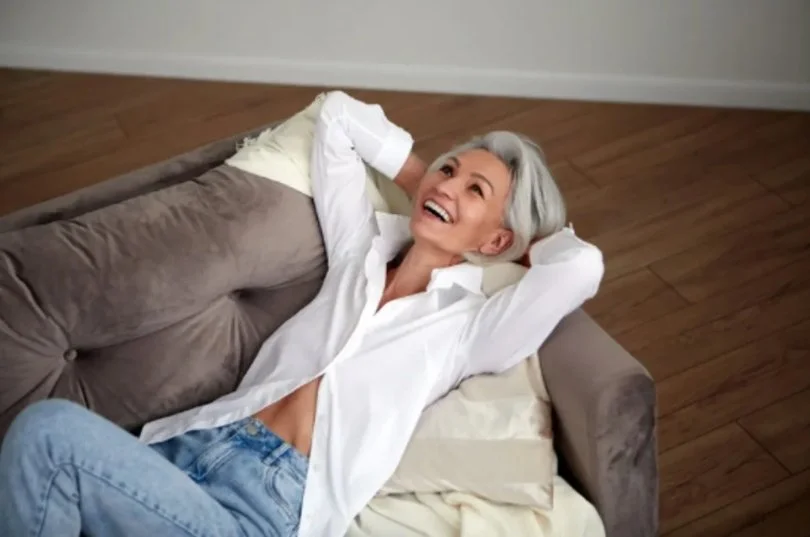 Smiling woman with gray hair lying on a couch with her hands behind her head, relaxed and happy.