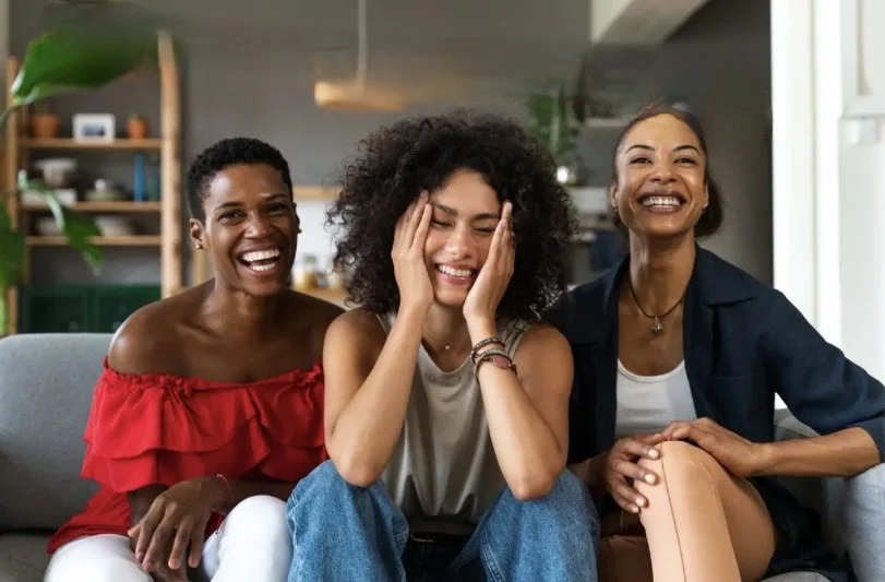 Three women sitting on a gray couch, smiling and laughing, in a cozy living room with wooden shelves and houseplants in the background.