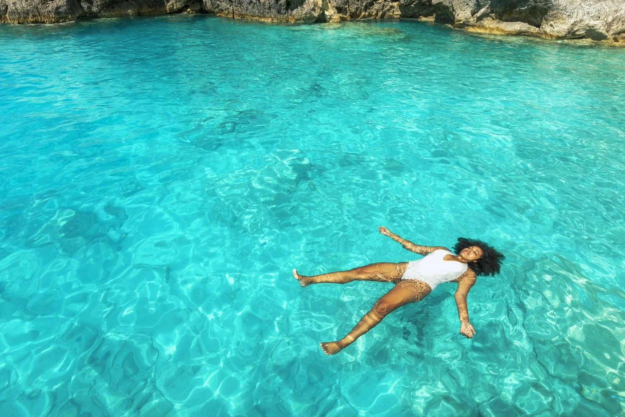 A woman in a white swimsuit relaxing and floating on her back in a clear blue lake surrounded by rocky cliffs.