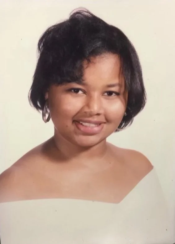A young woman with short black curly hair, wearing hoop earrings, smiling, against a plain light background.