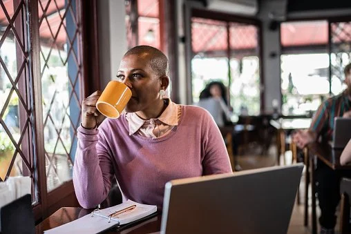 Woman with short hair drinking from a yellow mug inside a cafe, sitting at a table with a laptop and notebook.