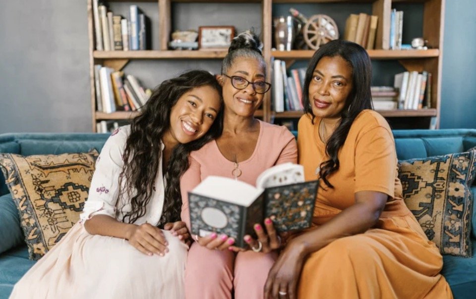 Three women sitting on a blue sofa, reading a book together, in a cozy living room with a bookshelf behind them.