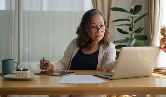 Older woman with glasses working on a laptop at a home desk, with a plant and sunlight in the background.