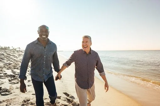 Two men walking hand-in-hand on a beach, smiling, with the ocean in the background during sunset.