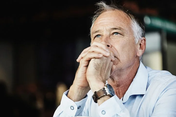 An older man with gray hair, wearing a white dress shirt, sits with his hands clasped near his mouth, looking thoughtful or pensive.