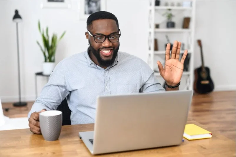 Smiling man with glasses waving at his laptop in a bright, modern home office with a coffee mug, a yellow notepad, and shelves with books and decor in the background.