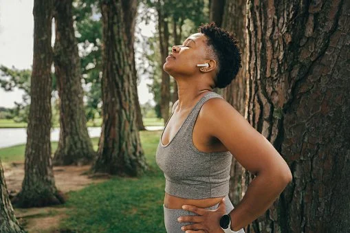 Woman stretching outdoors in a park with tall trees, wearing a gray sports bra and exercising.
