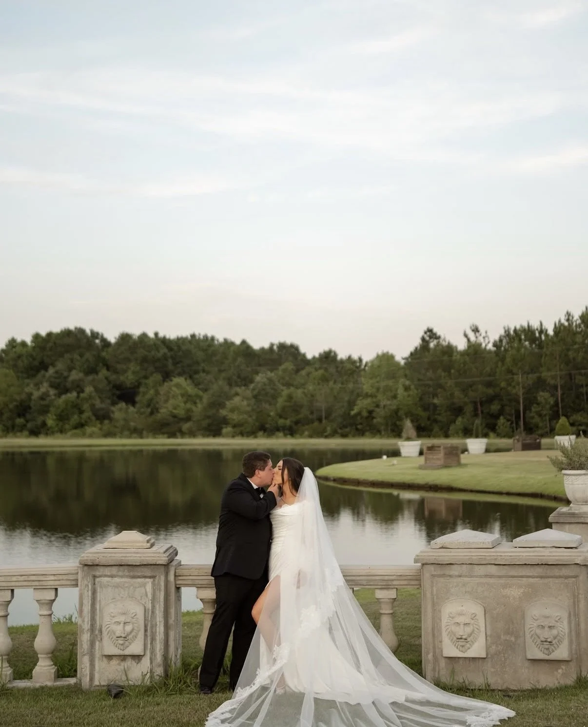 A newlywed couple sharing a kiss by a lake with a stone balustrade and decorative lion face carvings, surrounded by greenery and large white planters.