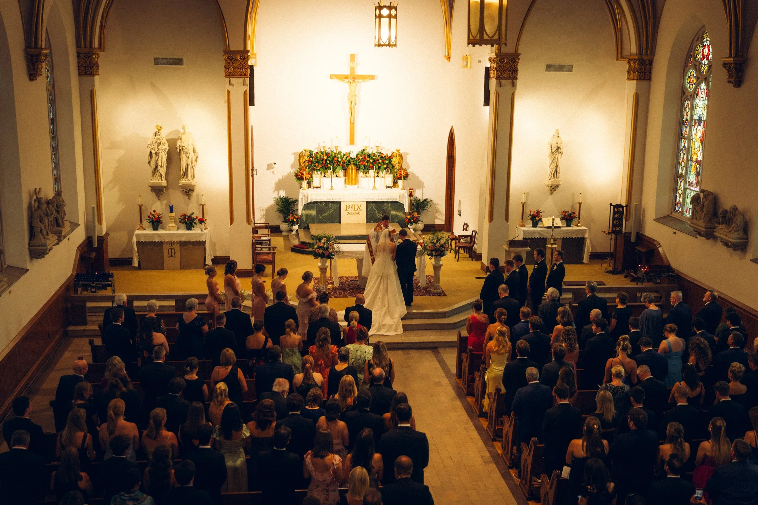A wedding ceremony taking place inside a church with a congregation gathered. The bride and groom are at the altar with a priest officiating. The church features religious statues, stained glass windows, and floral decorations.