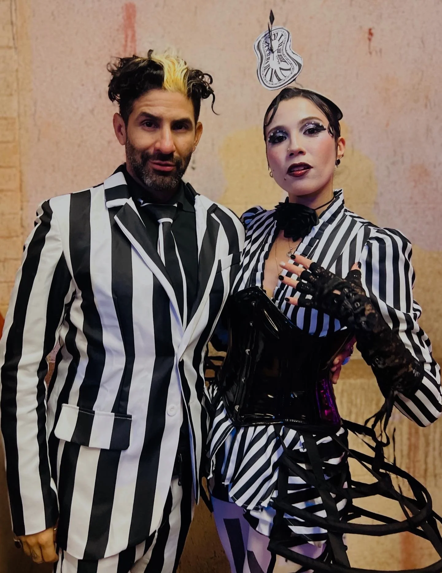 Two people dressed in black and white striped outfits, standing together indoors against a beige wall, with one person wearing a tall, melting clock hat.