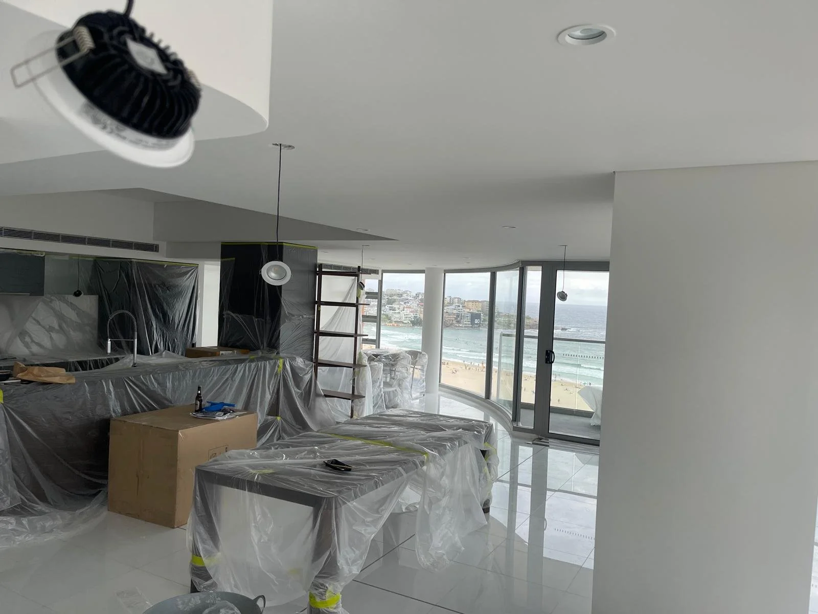 Living room under renovation with furniture covered in plastic, a ladder, and large floor-to-ceiling windows overlooking the beach and ocean.