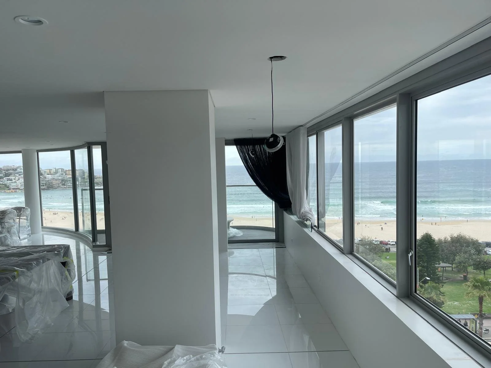 Interior of a high-rise apartment with large windows overlooking a beach and ocean, with some furniture and curtains still being installed.
