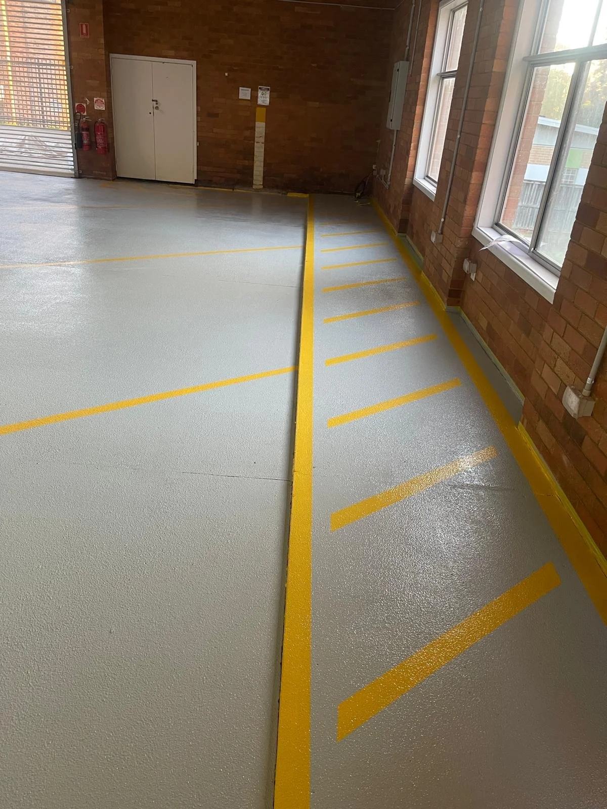 Empty indoor parking space with yellow lines marking parking spots and a yellow border along the wall. Brick walls and large windows letting in natural light.