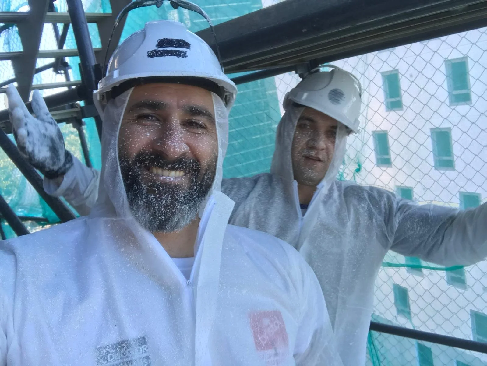 Two men in protective gear smiling and covered in paint on a construction site with scaffolding and a building with windows in the background.