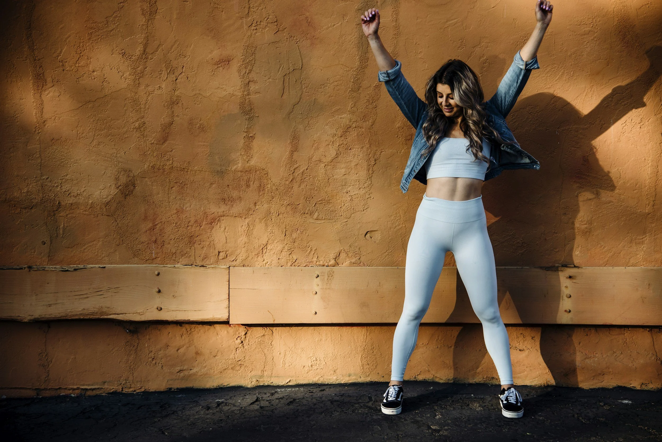 A woman in athletic wear standing against an orange textured wall with her arms raised and a joyful expression.