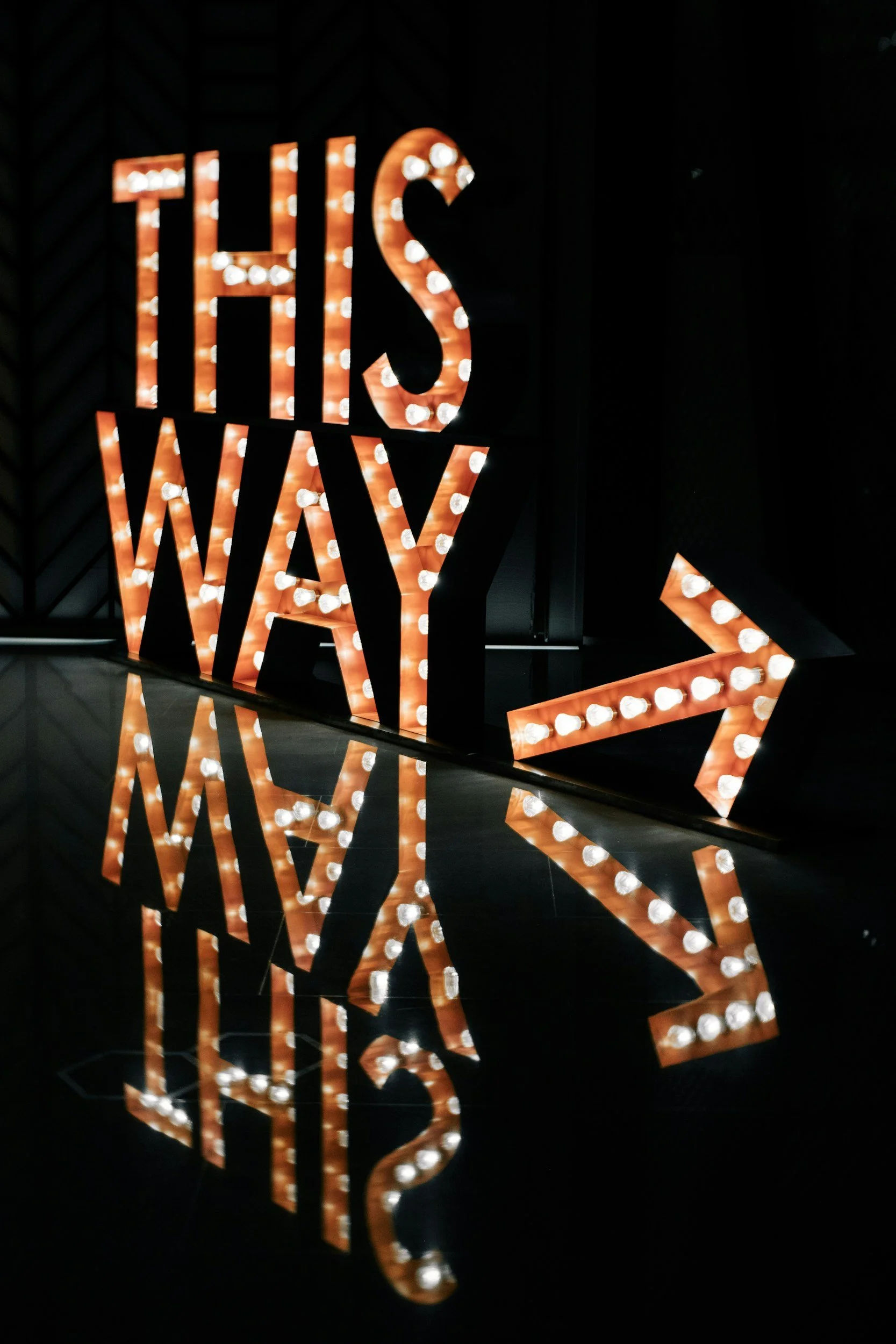 Decorative sign that says 'This Way' with an arrow, illuminated with light bulbs, and its reflection visible on the dark surface below.