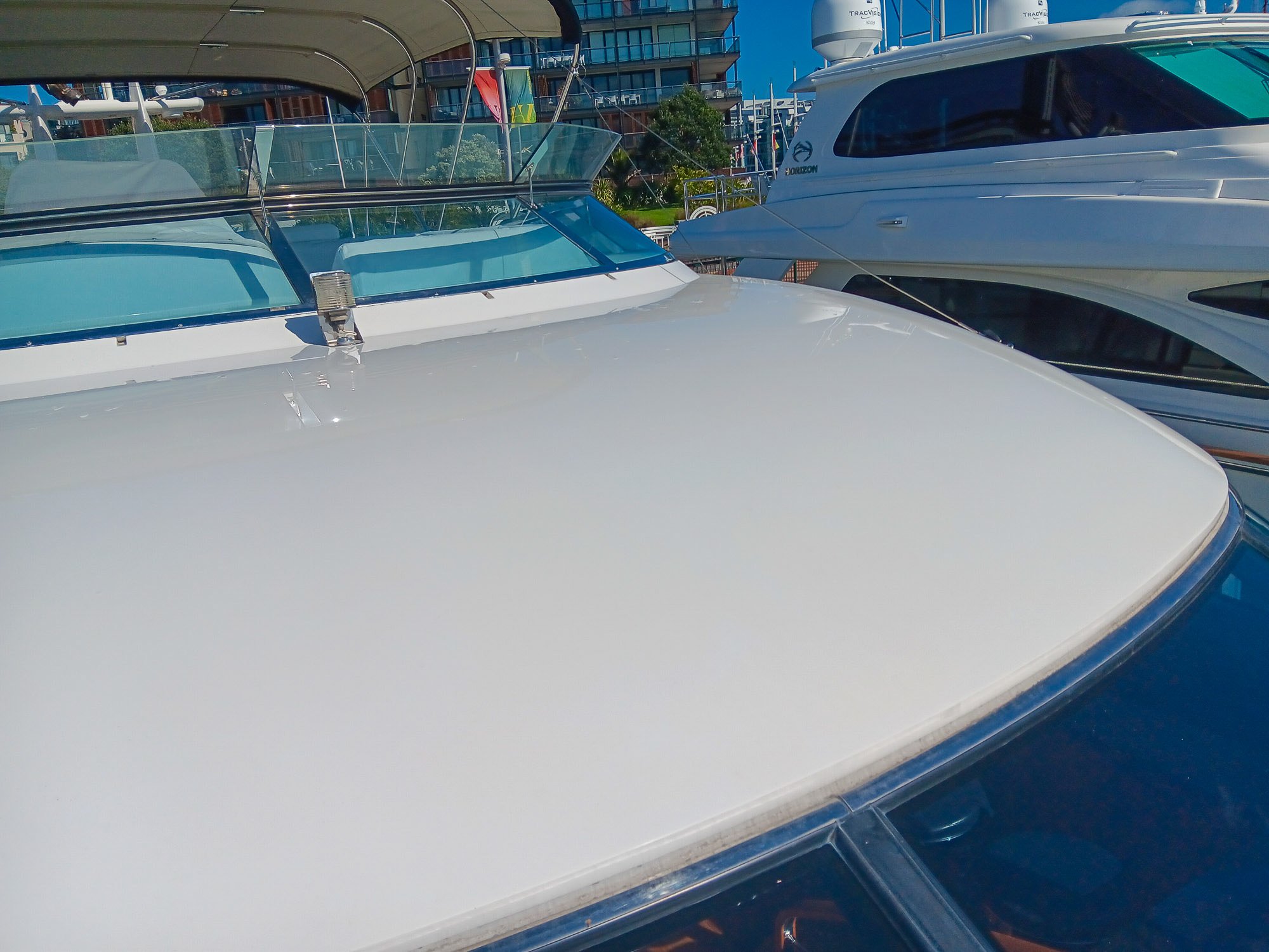 Close-up of the top of a white yacht docked at a marina, with other boats and modern buildings visible in the background.