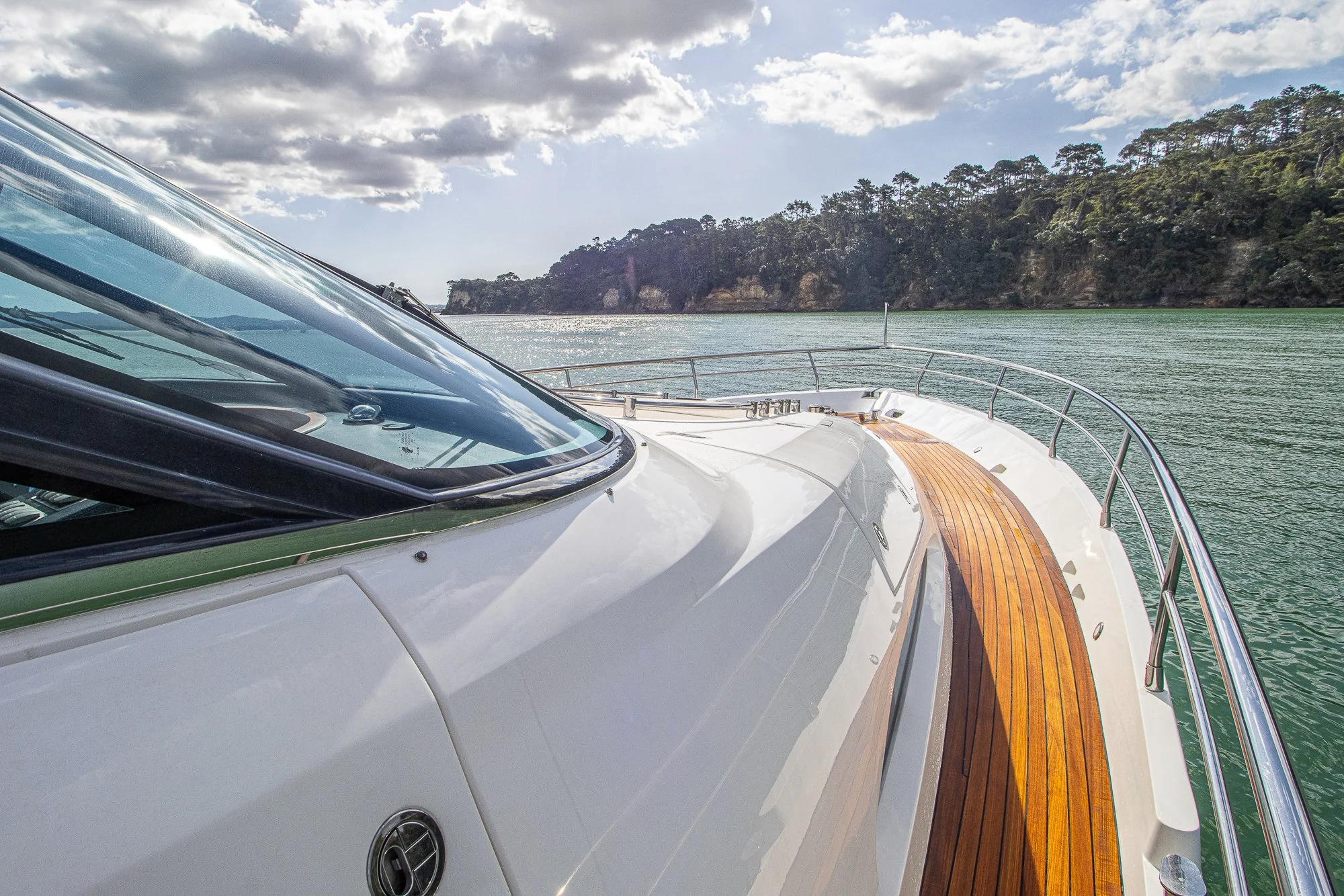 View from a yacht showing the deck with wooden flooring, railing, and a view of water and a tree-lined shoreline.