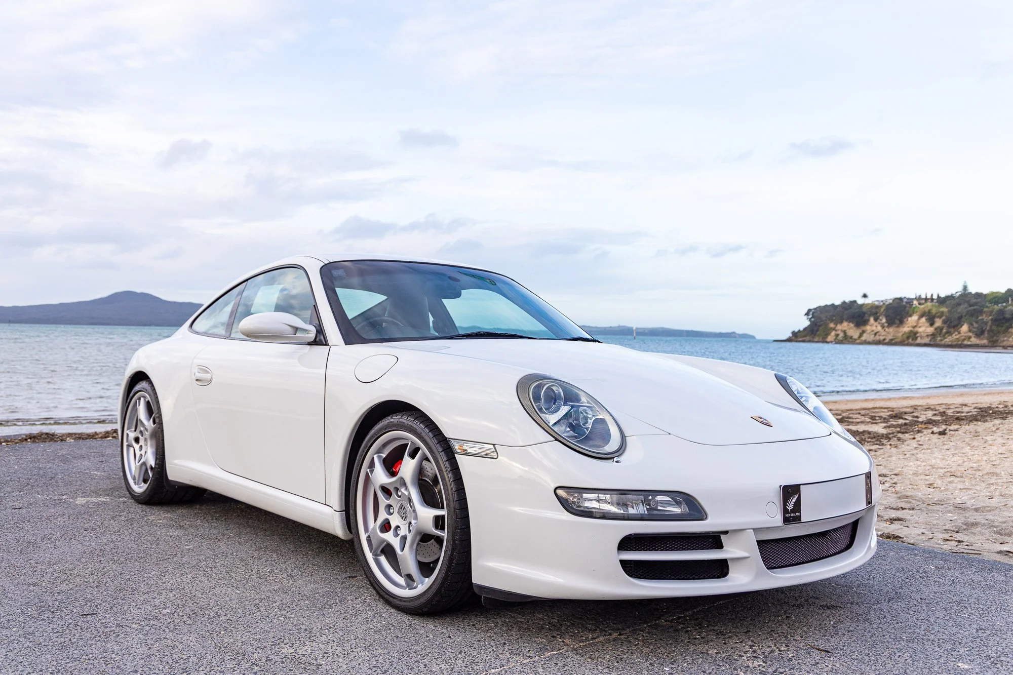 A white Porsche sports car parked on a paved surface near a beach, with water and a small island or landmass in the background on a partly cloudy day.