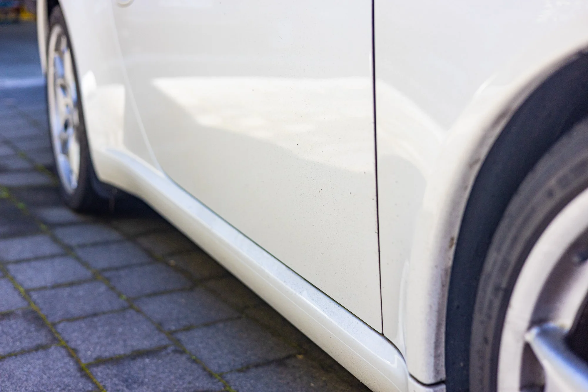 Close-up of dirty Porsche side, showing the bottom edge and part of the wheel, parked on a brick pavement.