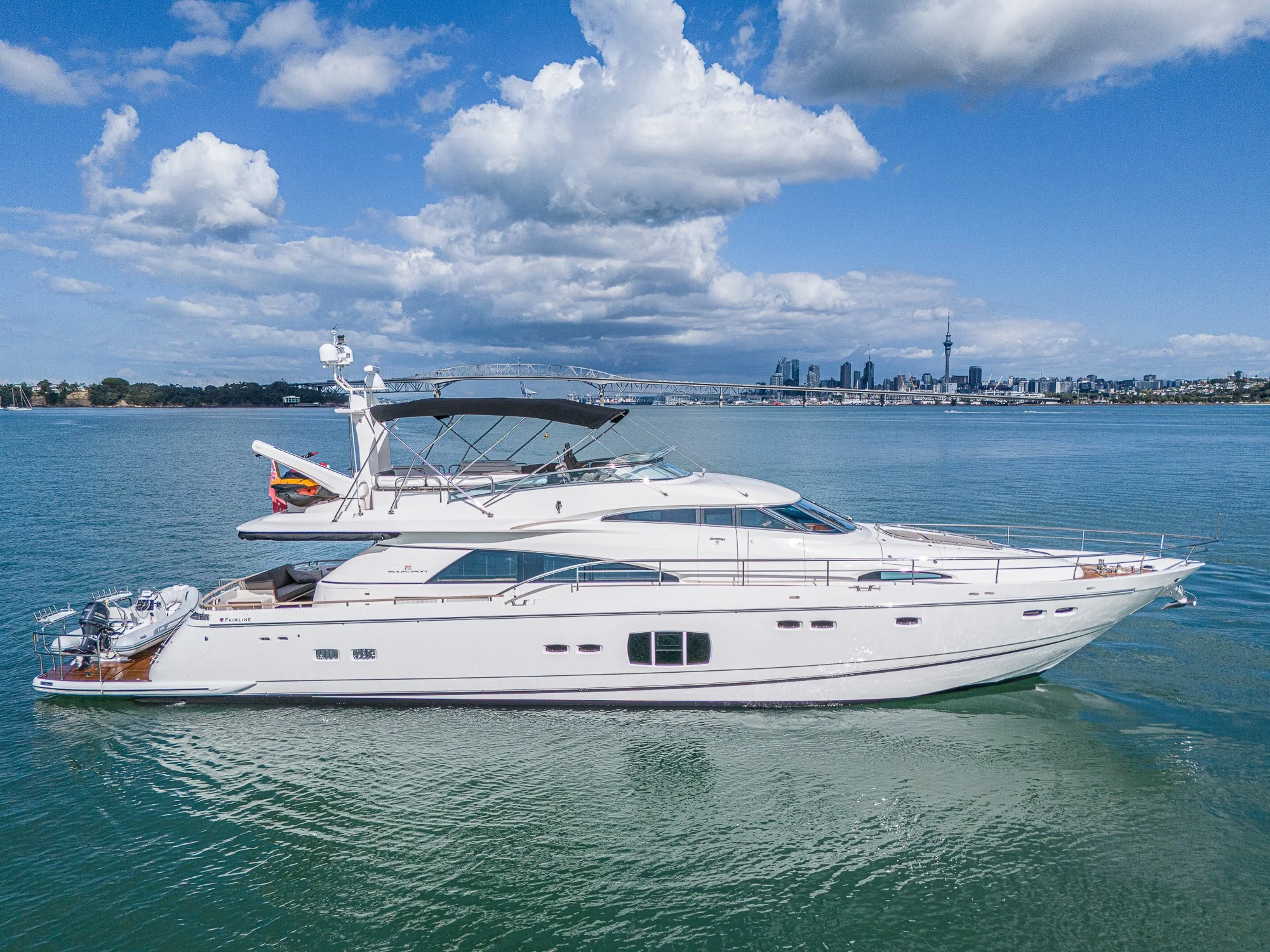 A white yacht cruising on water with city skyline and a bridge in background under cloudy sky.