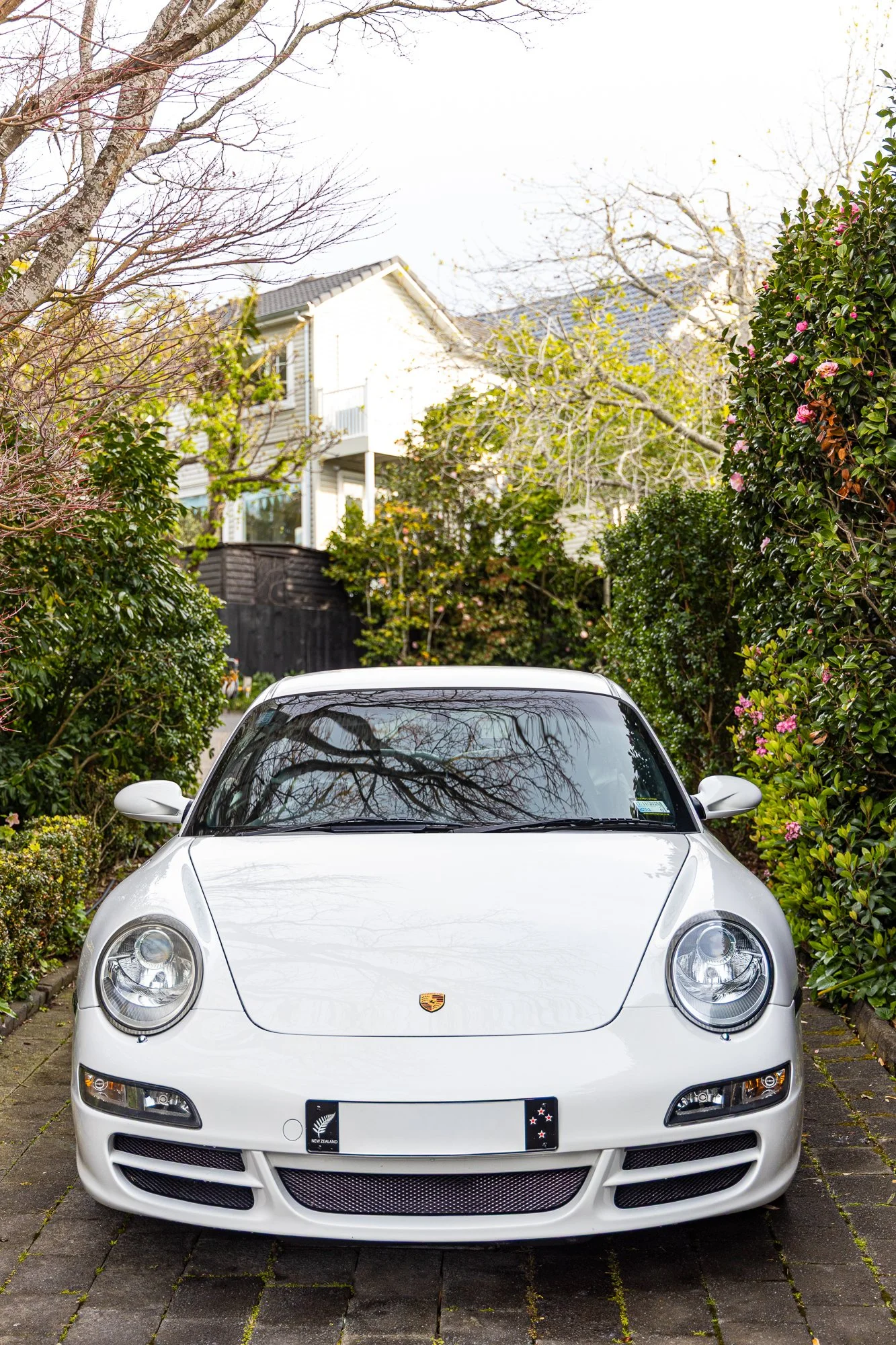 A white Porsche sports car parked on a brick driveway, surrounded by green bushes and trees with some pink flowers, in front of a residential house.