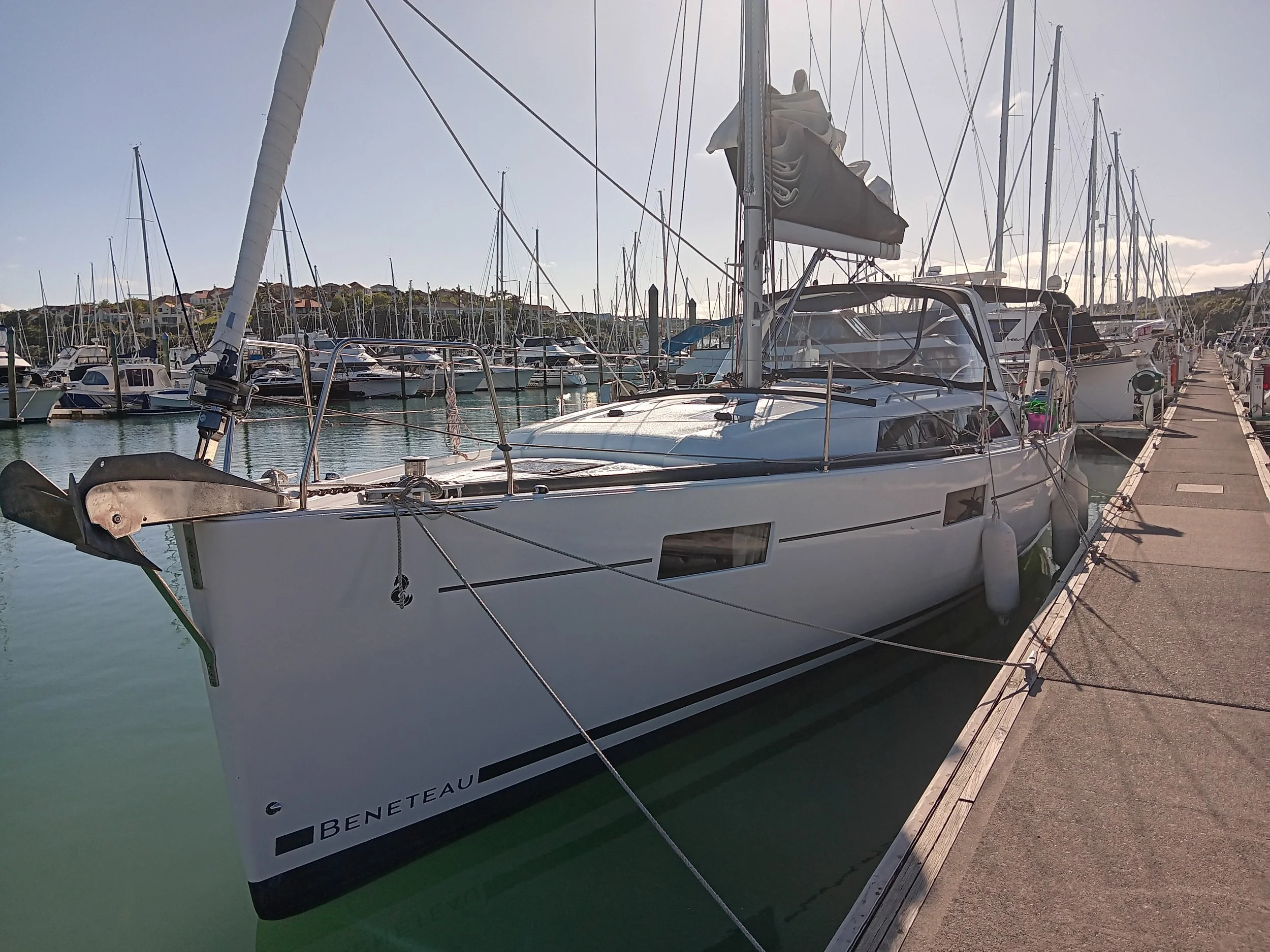 A white sailboat named Beneteau docked at a marina with other boats, on a sunny day with clear sky.
