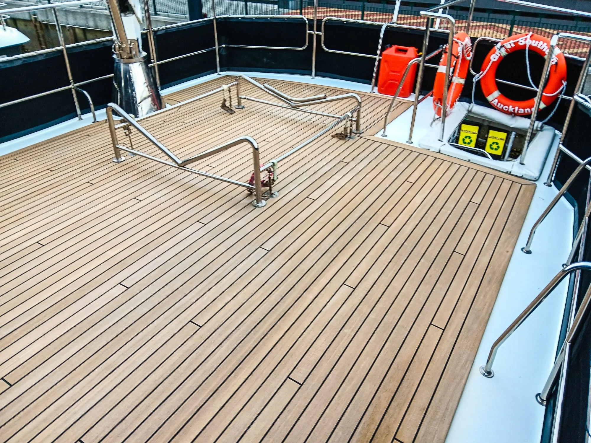 View of the deck on a boat, featuring wooden flooring, safety railings, life rings, safety equipment, and recycling bins.