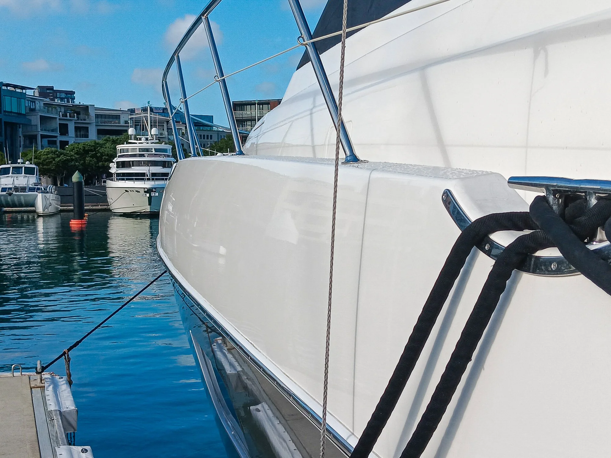 Close-up view of a white yacht moored at a marina with other boats and modern buildings in the background.