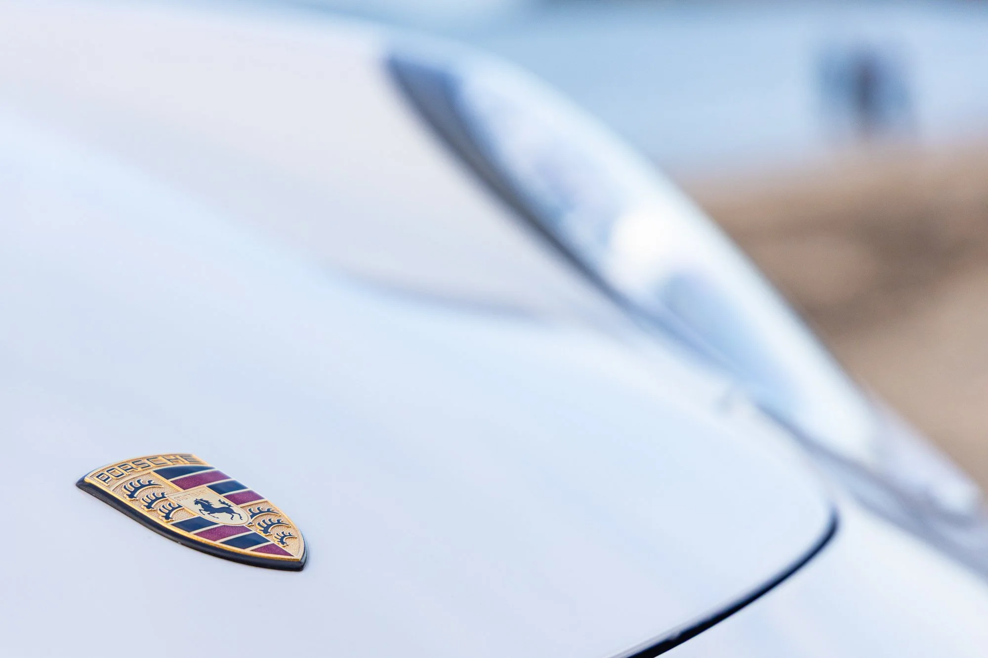 Close-up of a Porsche emblem on a white car hood with a blurred reflection in the background.