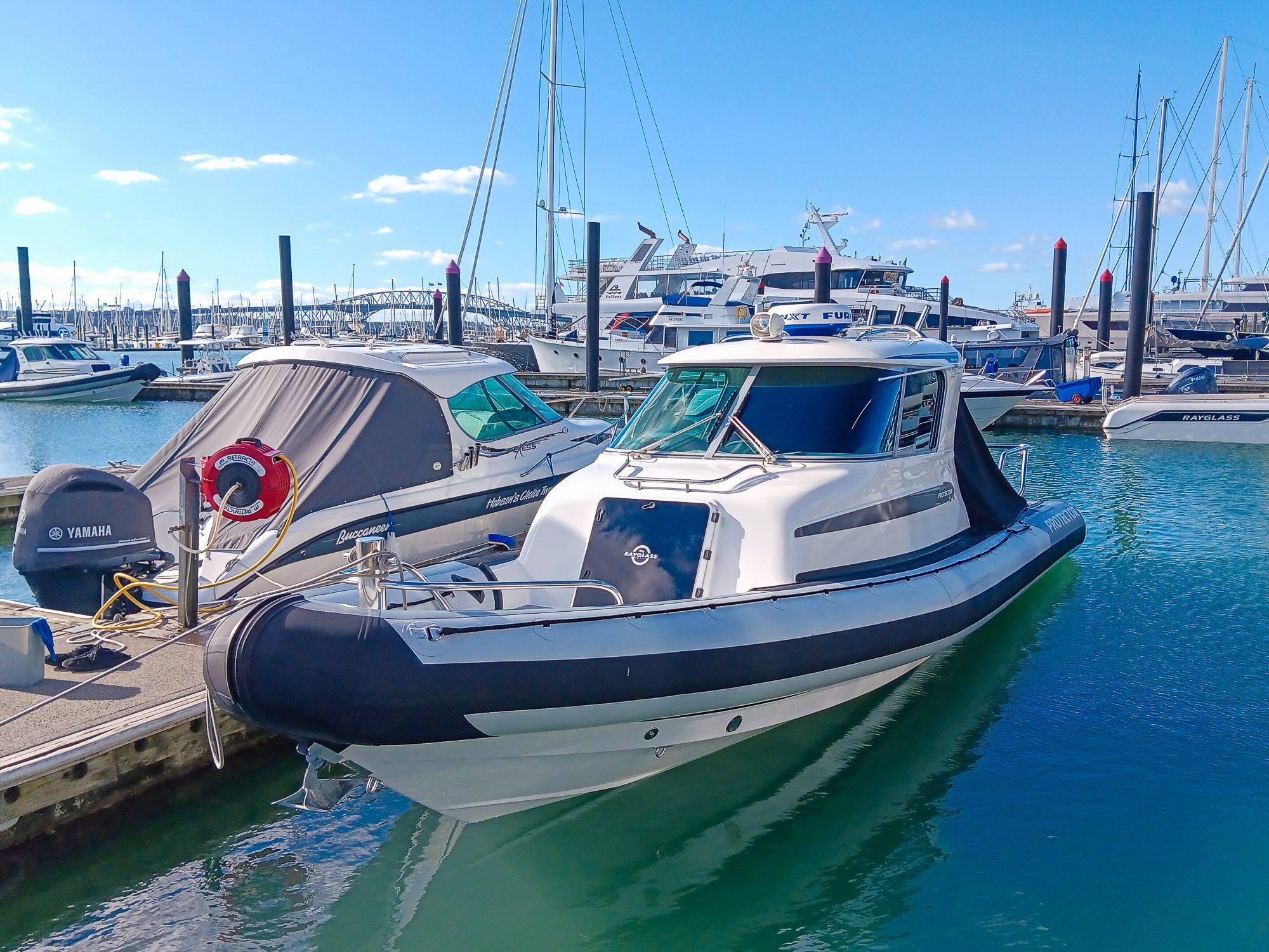 A white and black motorboat docked at a marina with several larger yachts and sailboats in the background, under a blue sky with scattered clouds.