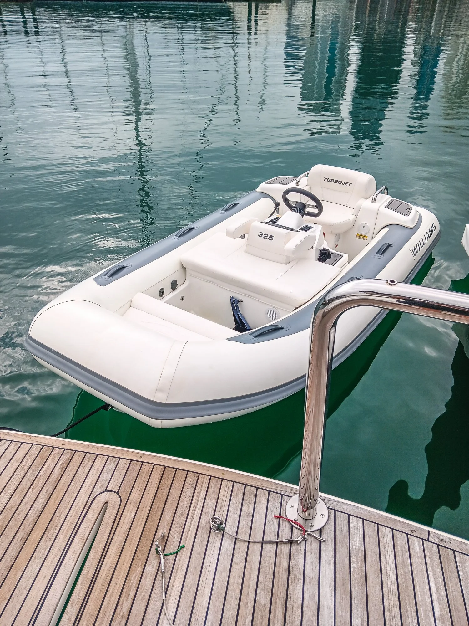 Small white inflatable boat docked at a wooden pier with a metal railing, on calm water with reflections of nearby boats or structures.