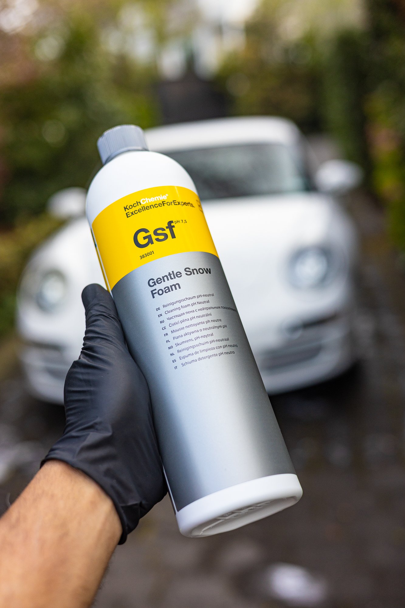 A person wearing a black glove holding a bottle of gentle snow foam car wash in front of a white car with round headlights, outdoors with blurred trees in the background.