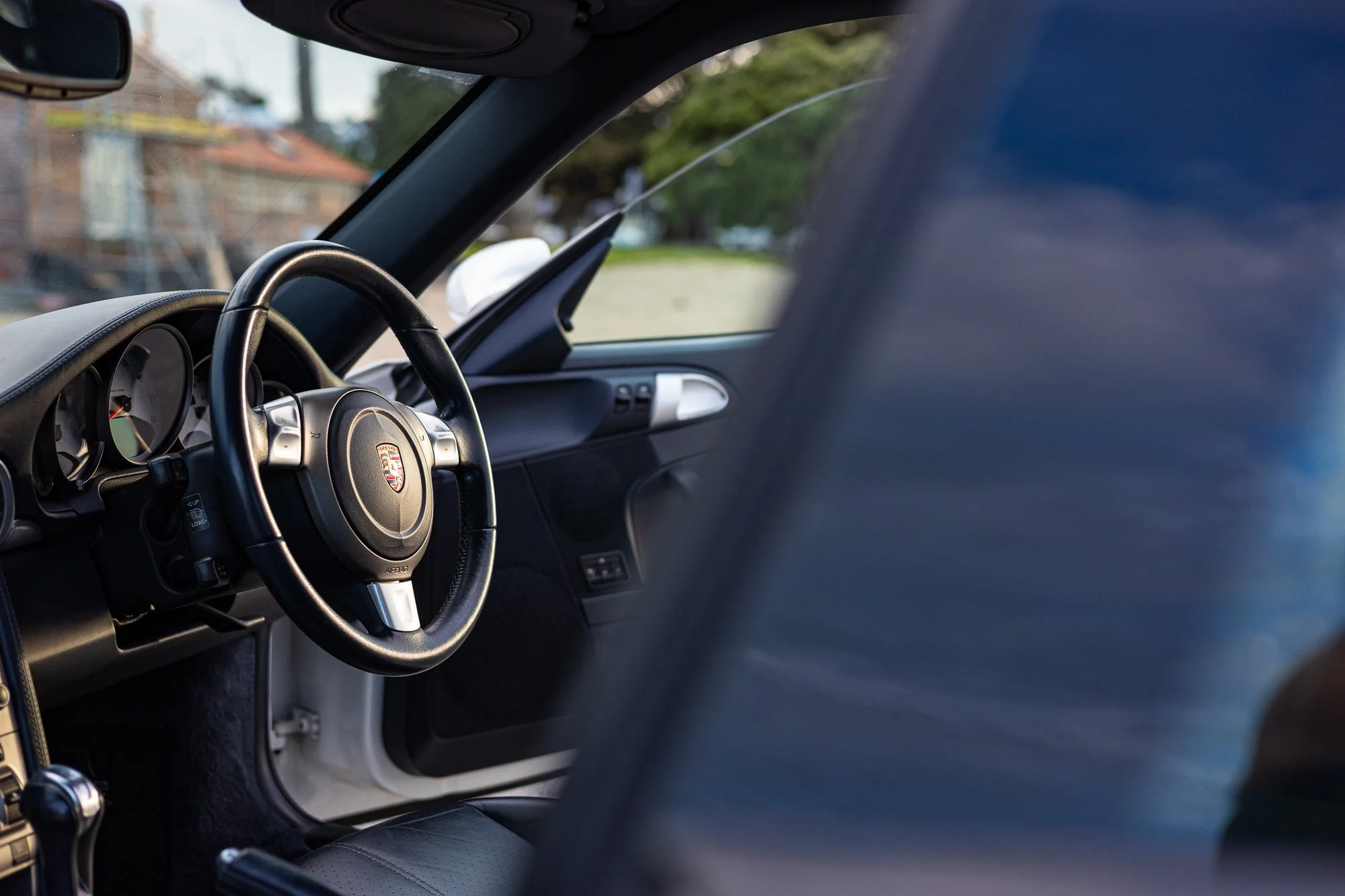 Interior view of a Porsche car with a black steering wheel and dashboard, partially obscured seat in the foreground, outside scenery visible through the window.