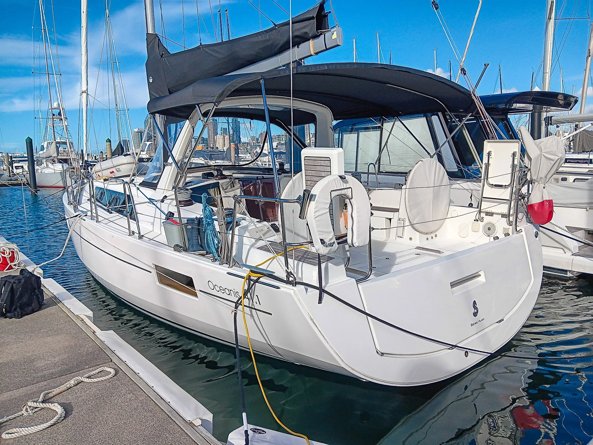A white motor yacht named Oceanis 41.1 is docked at a marina, with several sailboats and a city skyline in the background.