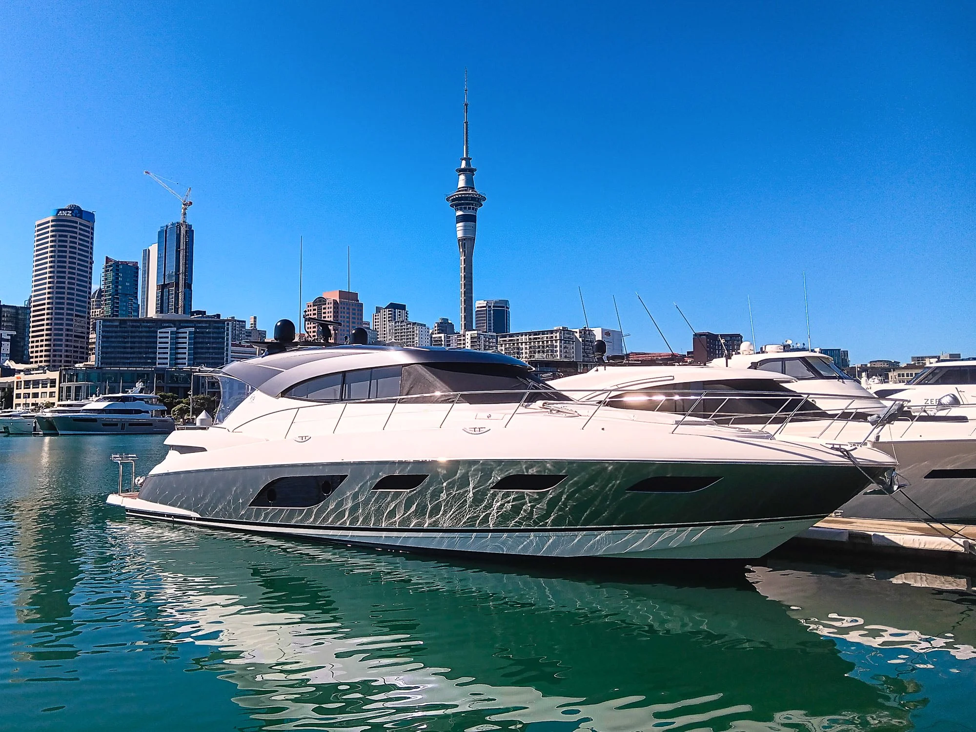 Luxury yachts docked at a marina with a city skyline including a tall tower in the background on a sunny day.