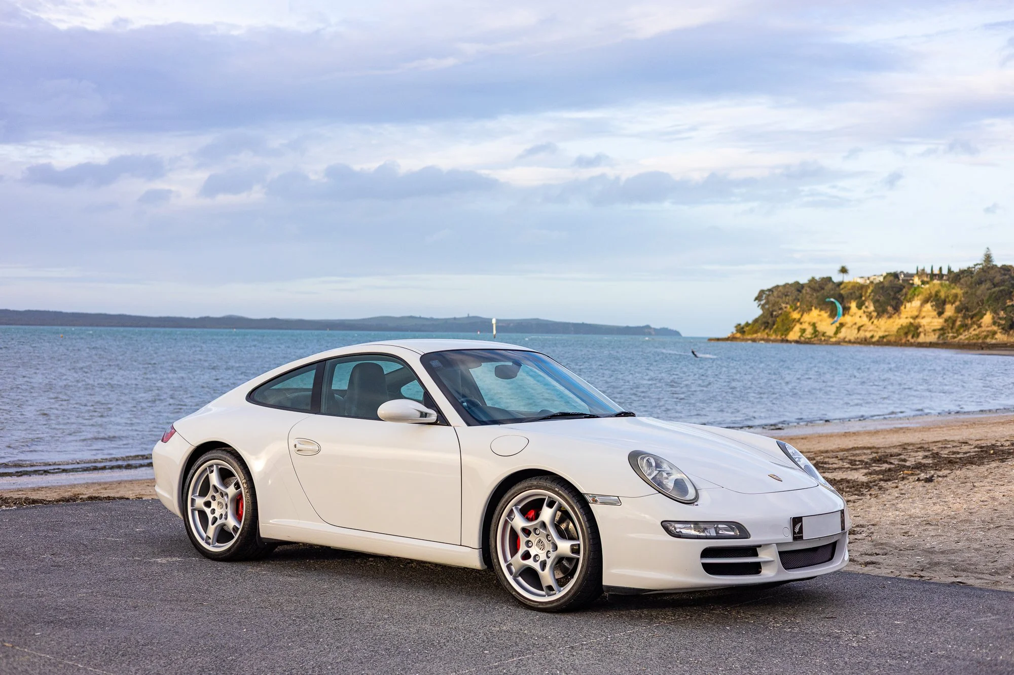White Porsche sports car parked near a beach with water and coastline in the background during daytime.