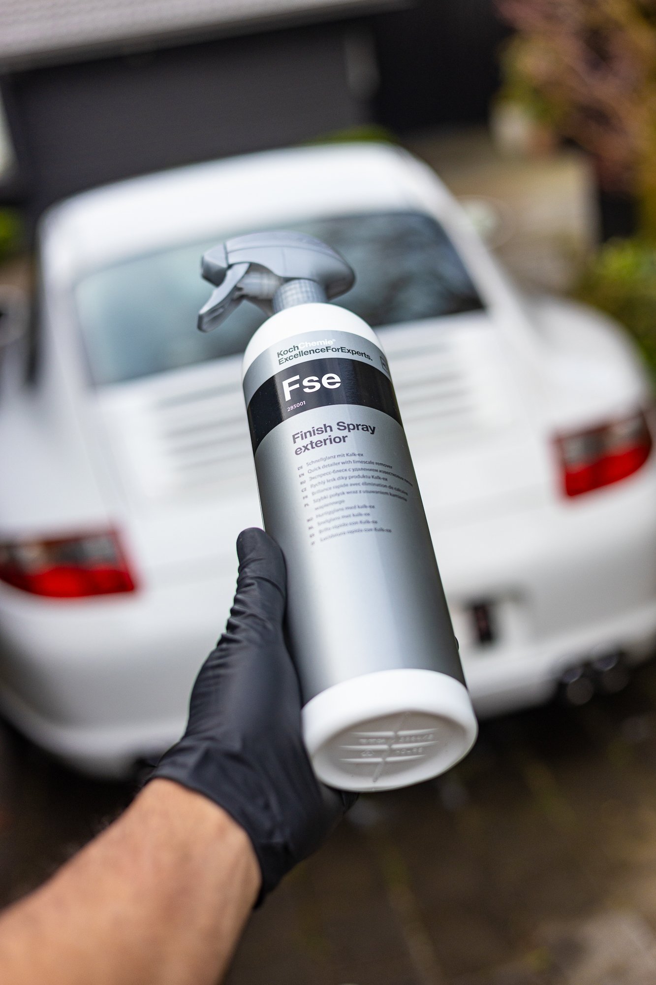 Person wearing black glove holding a spray bottle labeled 'Fse Finish Spray exterior' in front of a white classic car.