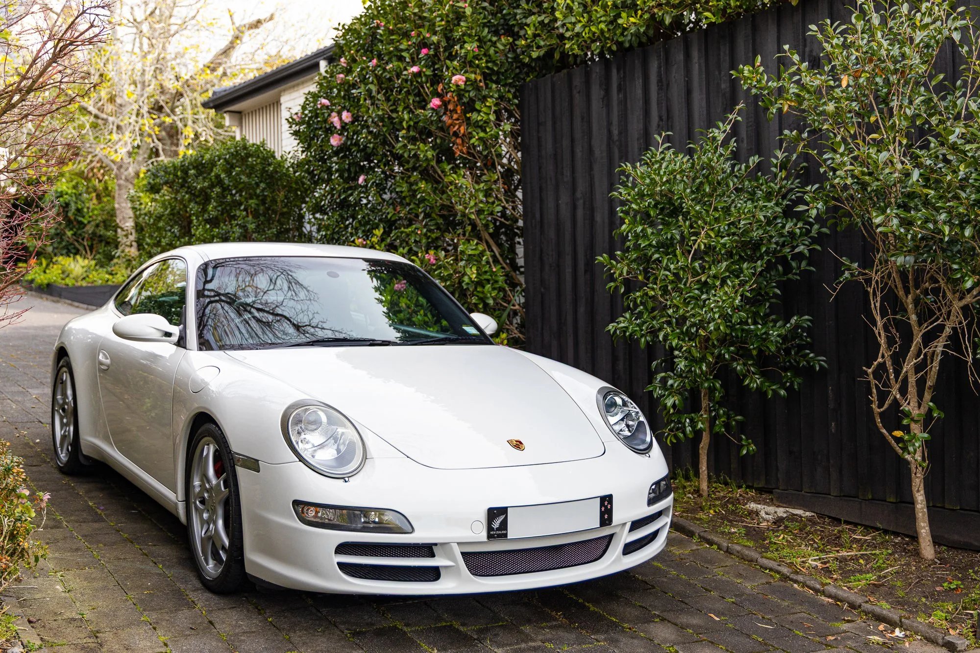 A white Porsche sports car parked on a brick driveway next to a black wooden fence, with bushes and trees in the background.