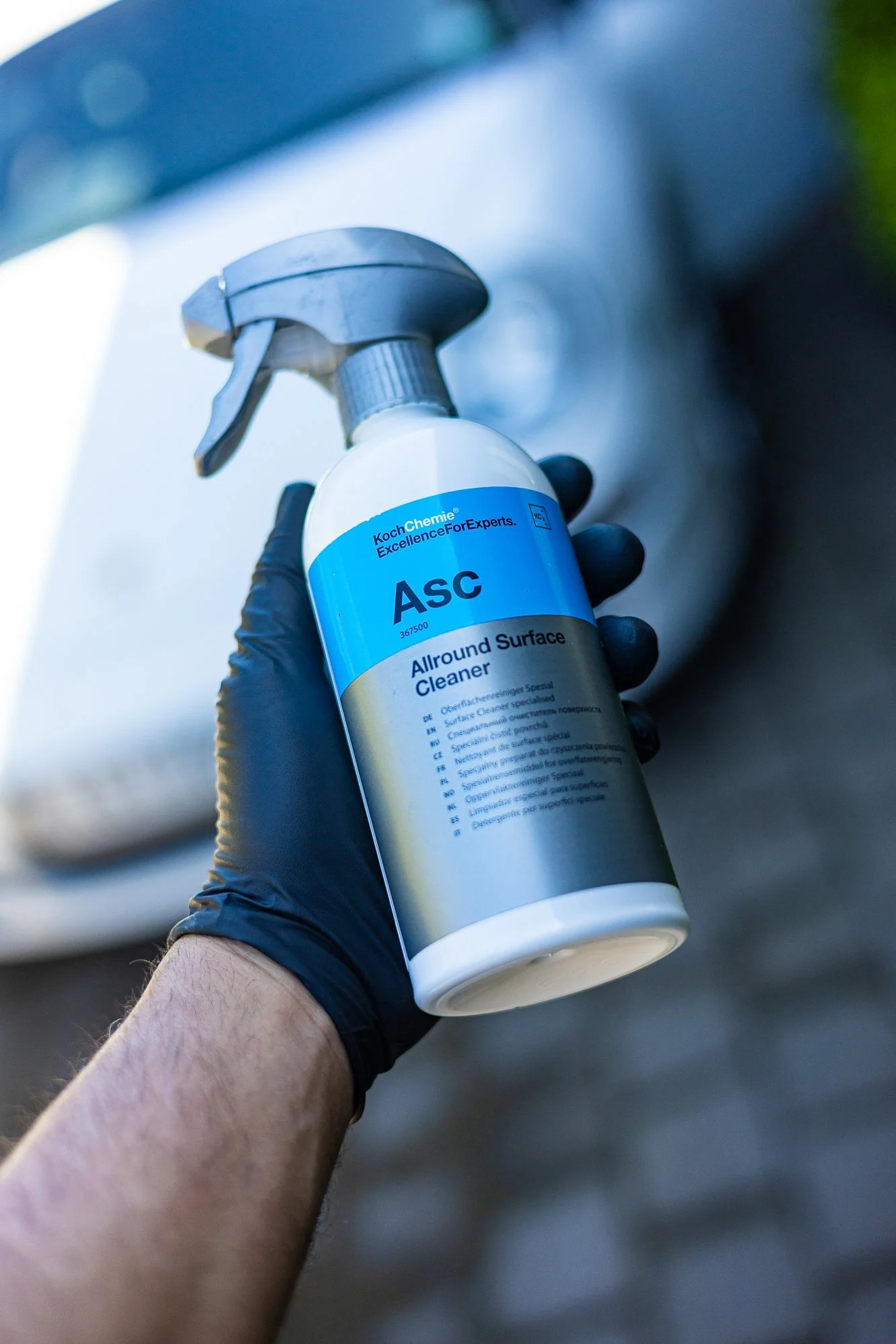 Person wearing a black glove holding a spray bottle labeled 'Allround Surface Cleaner' near a car, with the car's front visible in the background.
